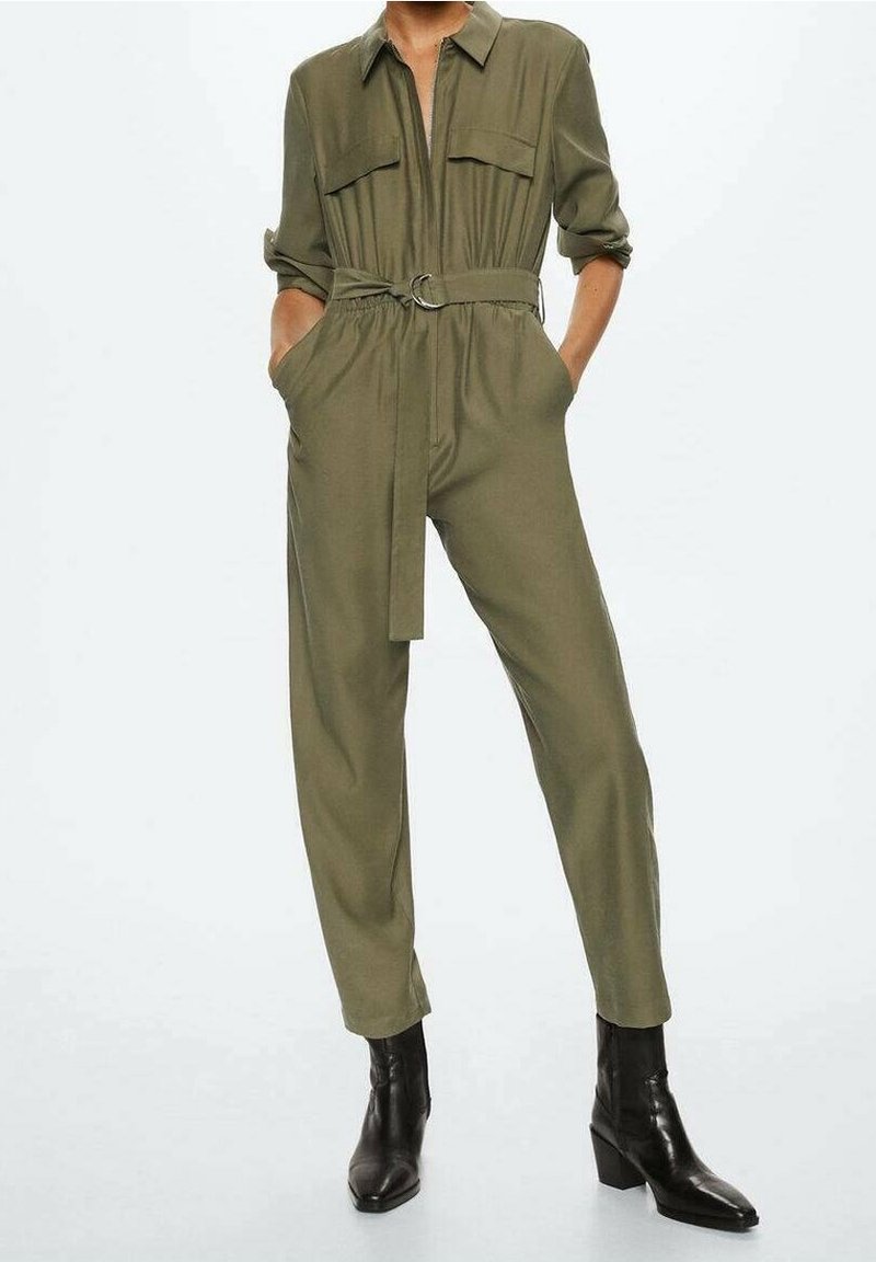 Femme portant une combinaison vert olive ceinturée avec manches retroussées et bottines noires, posant les mains dans les poches.