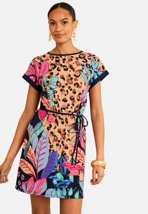 Žena, ktorá nosí krátke šaty s leopardím vzorom a farebnými tropickými listovými vzormi, zviazané v páse, doplnená veľkými zlatými náušnicami a náramkom.