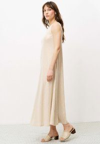 Ärmelloses, bodenlanges Kleid in hellbeige mit einem strukturierten, gerippten Muster. Kombiniert mit hellbeigen Rutsch-Schuhen, die einen dicken Absatz haben.