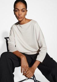 Frau mit zurückgebundenem Haar, die ein lockeres weißes Oberteil und schwarze Hosen trägt, sitzt auf einem Stuhl mit Metallrahmen vor einem weißen Hintergrund.