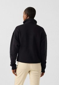Pull en polaire noir avec un col haut, coupe décontractée et épaules tombantes, associé à un pantalon ajusté beige clair. Texture douce.
