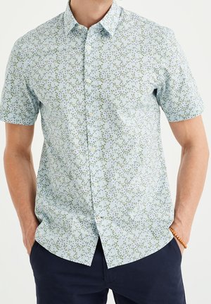 Mann trägt ein hellblaues kurzärmliges Hemd mit kleinem grünem und lila Blumenmuster und dunkle Hose, Hände in den Taschen.