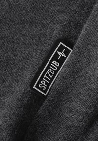 Dunkelgrauer Stoff mit einer weichen Textur, der ein schwarzes Etikett mit der Aufschrift 'SPITZBUB' und einem grafischen Designelement zeigt.