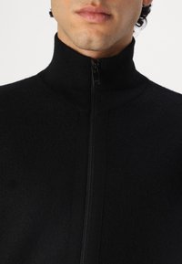 Maglione in pile zip-up nero con colletto alto, caratterizzato da un tessuto testurizzato e una chiusura a cerniera liscia sul davanti.