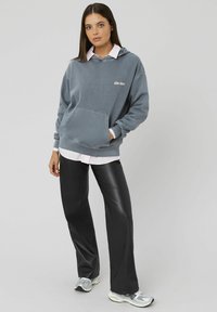 Haut en coton gris avec le texte "Soyez gentil", poche avant et capuche avec cordon de serrage, superposé sur une chemise à col rose. Pantalon en simili cuir noir.
