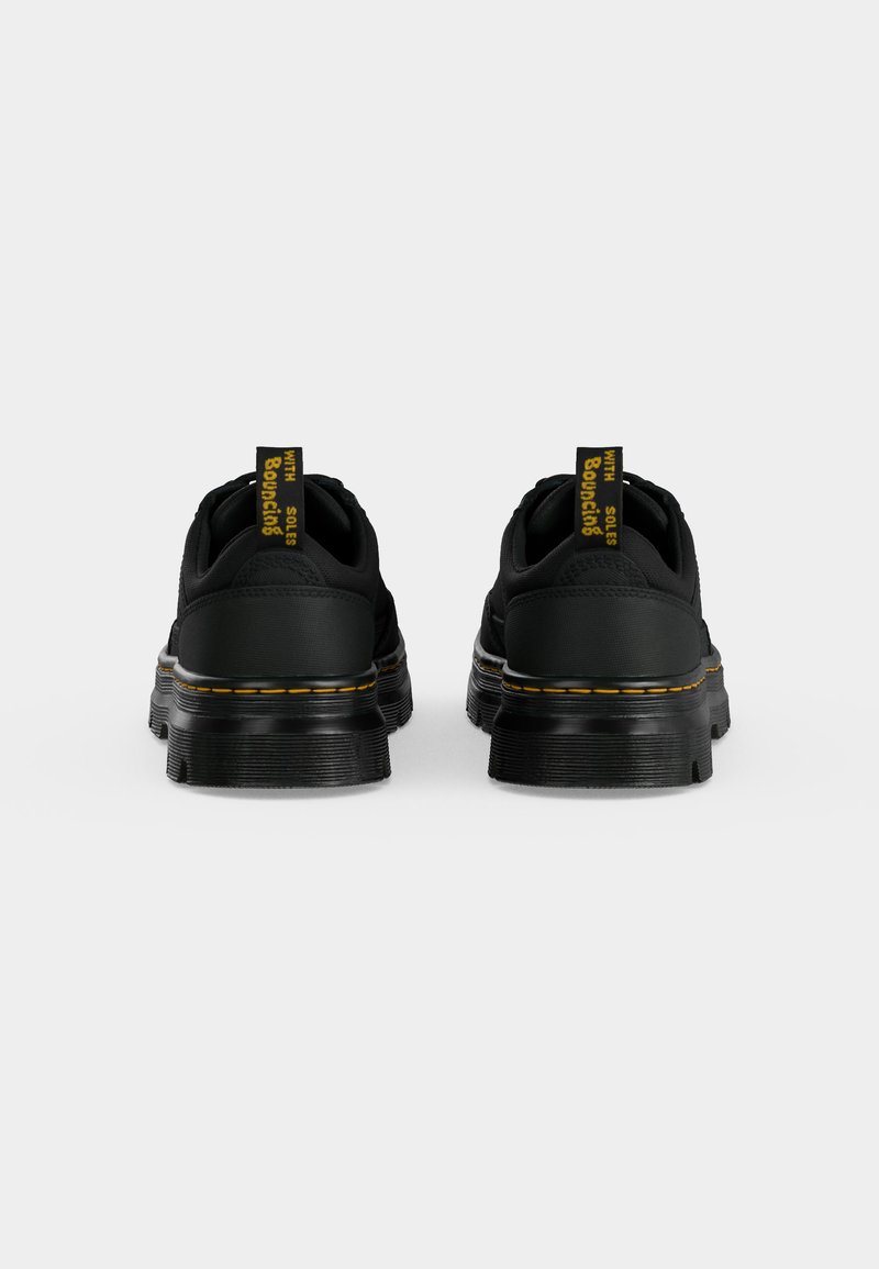 Paar schwarze flach geschnittene Schuhe mit dicken Sohlen, gelben Nähten und Fersenschlaufen mit dem Schriftzug "WITH Bouncing SOLES", von hinten auf weißem Hintergrund gezeigt.