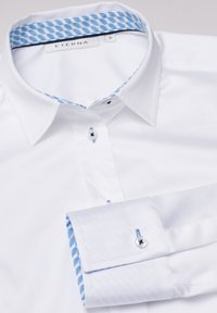 Chemise blanche avec un col à motifs bleus et des coutures d'accent bleu. Comprend des boutons noirs et une texture lisse et tissée. Étiquette ETERNA visible.