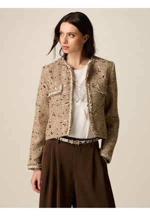 Kobieta w beżowej, cekinowej krótkiej kurtce na białej koszulce z nadrukiem, brązowe spodnie z szerokimi nogawkami i wysokim stanem oraz wzorzysty pasek, patrząca w lewo.