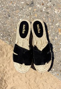 Paire de sandales tissées beige avec des lanières en daim noir sur une surface texturée en pierre et sable, nom de marque "sapha" visible sur les semelles intérieures.