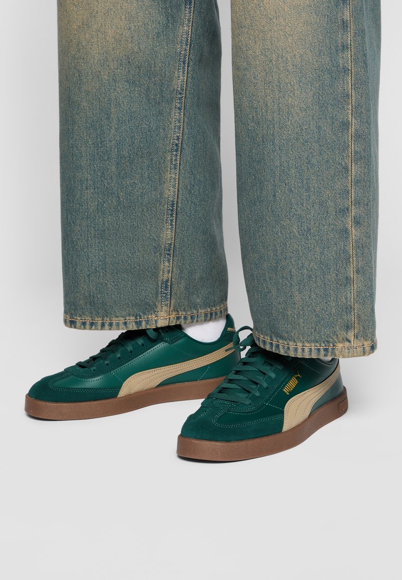 Grüne Wildleder- und synthetische Sneakers mit beigen Akzenten, ausgestattet mit einer Gummisohle und runden Schnürsenkeln, kombiniert mit ausgewaschenen blauen weit geschnittenen Jeans.