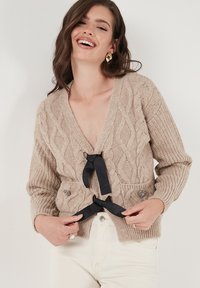 Beige knitted cardigan with cable patterns, v-neck, black ribbon tie, and decorative pockets featuring embellishments.