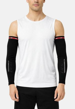 Uomo con maglia bianca senza maniche e manicotti neri con bande di logo rosse e grigie, in piedi con le braccia rilassate ai lati.