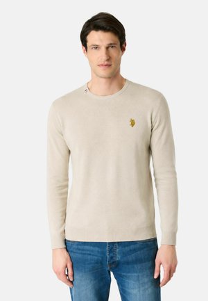 Uomo che indossa un maglione beige a maniche lunghe con un piccolo logo ricamato sul petto e jeans blu, in piedi davanti a uno sfondo uniforme.