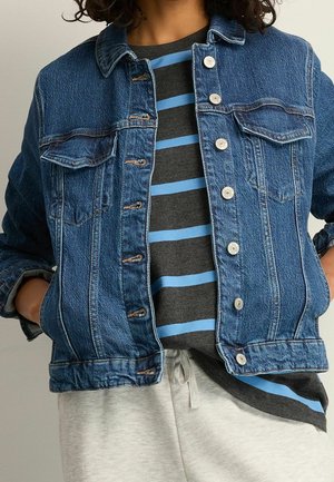 Personne portant une veste en jean bleue sur une chemise grise avec des rayures horizontales bleu clair et un pantalon gris clair avec une taille à cordon.