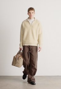 Béžový sveter s textúrou a výstrihom do V, doplnený voľnými hnedými nohavicami. Nesie koženú tašku v odtieňoch hnedej a tan, na nohách čierne topánky.