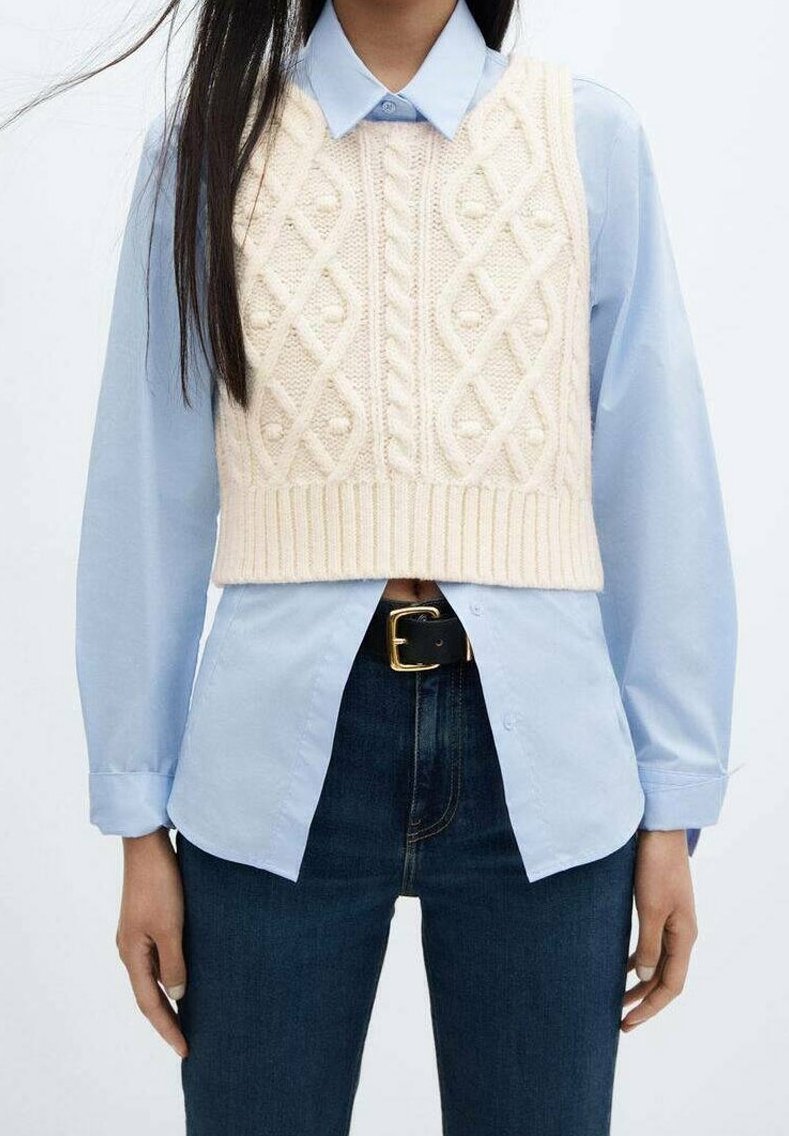 Femme portant un pull sans manches en tricot torsadé crème par-dessus une chemise bleue claire à boutons, un jean foncé, et une ceinture noire avec une boucle dorée.
