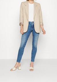 Kvinna som bär en beige blazer över en vit topp, tillsammans med blå skinny jeans och beige stilettklackar, stående mot en enfärgad bakgrund.