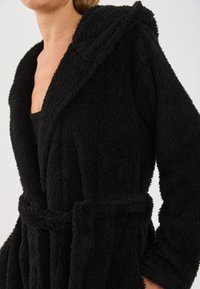 Rochie de baie neagră pufoasă cu glugă, având un braț cu curea și mâneci lungi. Țesătura are o suprafață moale și texturată.