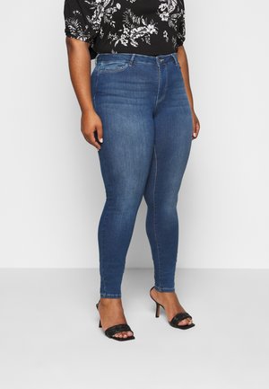 Kvinna som bär blå smala jeans, svart blommig blus och svarta högklackade öppna sandaler står mot en enfärgad bakgrund.