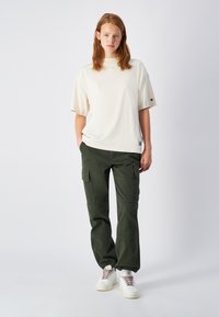 Camiseta de algodón crema con mangas cortas, combinada con pantalones cargo verde oscuro que tienen bolsillos y puños elásticos. Lleva zapatillas blancas.
