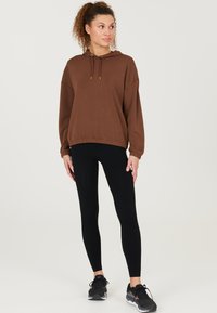 Haut à capuche surdimensionné marron avec cordon de serrage, leggings noirs et chaussures de sport noires. Tissu lisse, coupe décontractée, design minimaliste.