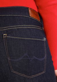 Jeans de mezclilla azul oscuro con costuras naranjas, una etiqueta de cuero en la cinturilla y un bolsillo trasero con un detalle de bordado en forma de ola.