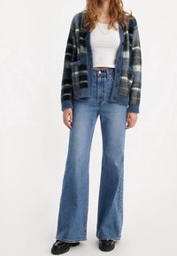 Cardigan à carreaux bleu marine et gris avec une texture douce, doté de boutons et de poches, associé à un jean évasé bleu clair et des chaussures noires.