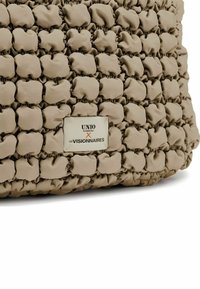 Sac beige avec un design texturé comportant des sections gonflées et froncées. Comprend une étiquette de marque indiquant "UNIO Hamburg x LES VISIONNAIRES."