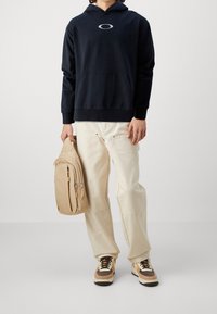 Sudadera con capucha azul marina con bolsillo delantero, mochila beige con cremalleras, pantalones color crema y zapatillas con detalles en marrón.