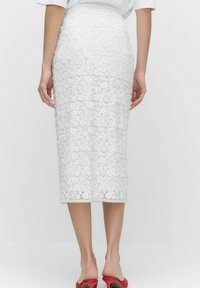 Jupe crayon en dentelle blanche avec un motif floral, longueur genou, accompagnée de chaussures ouvertes rouges.