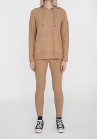 Maglione a coste color cammello con cappuccio e tasca frontale, abbinato a leggings a coste coordinati. Indossato con scarpe da ginnastica nere e bianche.