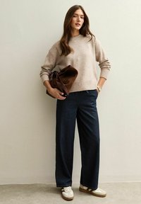 Suéter de punto beige, jeans de pierna ancha oscuros y zapatillas blancas con detalles marrones. Bolsa marrón y reloj visibles, cabello largo y ondulado.