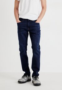 Jean en denim bleu foncé avec une coupe slim, cinq poches et des coutures orange. Associé à des chaussures grises texturées et un t-shirt blanc.