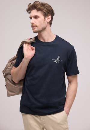 Mężczyzna w granatowej koszulce z małym białym wzorem liścia i współrzędnymi, trzymający beżową kurtkę na ramieniu, w beżowych spodniach, patrzący w lewo.