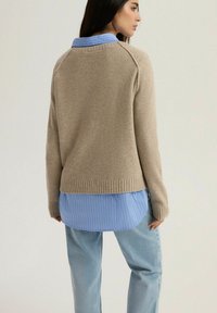 Beżowy sweter z dzianiny z prążkowanym dołem, noszony na jasnoniebieską koszulę w paski. Projekt charakteryzuje się okrągłym dekoltem i rękawami raglanowymi.