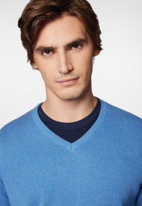 Jasnoniebieski sweter w serek uszyty z miękkiego, gładkiego materiału, założony na ciemnoniebieską koszulę. Tekstura wydaje się mieć drobny splot z czystym wykończeniem.