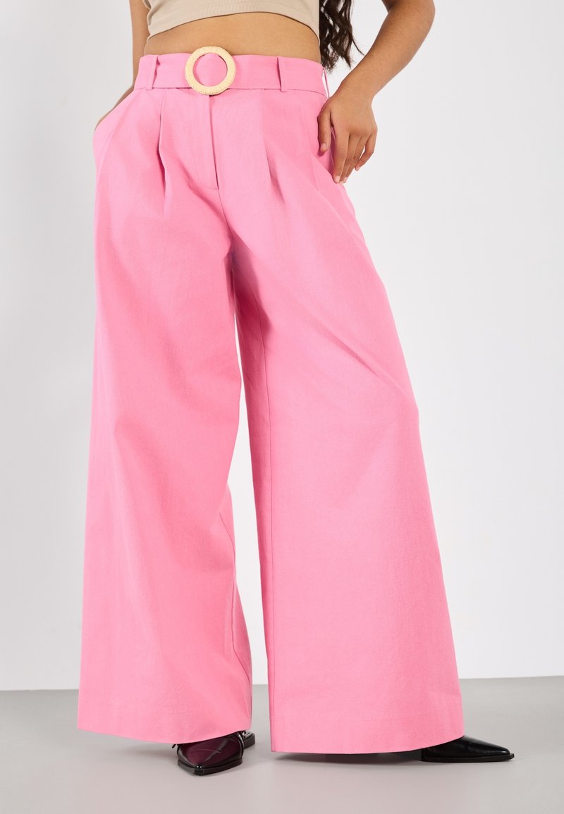 Femme portant un pantalon rose taille haute à jambes larges avec une boucle de ceinture circulaire et des chaussures noires pointues sur un fond uni.