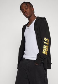 Sudadera con cremallera negra con el logo "BULLS" en dorado en la manga, llevada sobre una camiseta sin mangas blanca. Pantalones negros completan el atuendo.