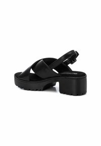 Sandalias negras con una suela plataforma gruesa, que presentan tiras cruzadas y una correa en el talón ajustable. Textura de cuero suave con un acabado mate.