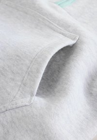 Világosszürke pulóveranyag finom textúrával, türkizkék díszítéssel és oldalzseb részlettel. Varrási varratok láthatók.