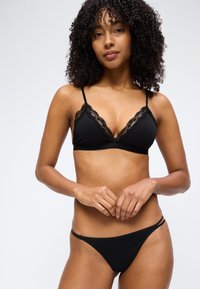 Γυναίκα με σγουρά μαλλιά που φοράει μπραλέτο με μαύρη δαντέλα και μαύρο εσώρουχο στυλ μπικίνι, σε απλό φωτεινό φόντο.