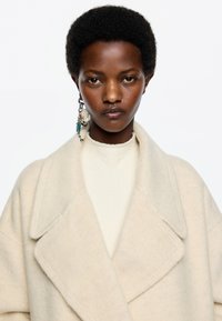 Cappotto oversize beige in lana con ampie rever, indossato sopra un dolcevita crema. Presenta un tessuto testurizzato e un orecchino di design con materiali misti.
