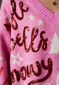 Pull rose avec des étoiles blanches et des sapins de Noël, avec une inscription en sequins rouges « elf walks snowly ».