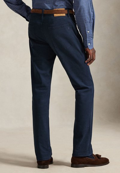 Calças de algodão azul-marinho com pernas direitas, dois bolsos traseiros e uma etiqueta de couro no cós. Sapatos de camurça complementam o conjunto.