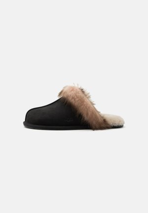 Sort slip-on hjemmesko med tyk tan og cremefarvet pelskant omkring hælen og indersiden, flad sål og UGG-logo på siden.