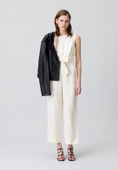 Mono marfil con cintura anudada, chaqueta de cuero negra caída sobre el hombro, y tacones negros con tiras y detalles dorados. Textura suave, corte de pierna amplia.