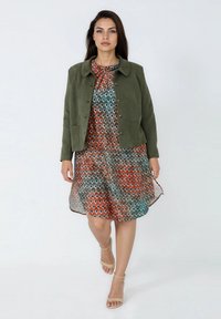 Olijfgroene suède jas over een meerkleurige, gestippelde jurk, met een ronde kraag en knoopsluiting, met een doorschijnende, vloeiende textuur.