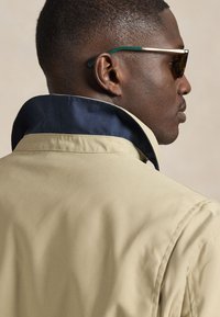 Giacca beige con colletto blu navy, caratterizzata da una texture liscia e una silhouette strutturata. Gli occhiali da sole hanno lenti scure con accenti verdi.