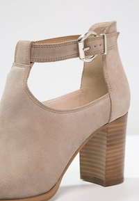 Beige mockasin-känga med medelhög blockklack, justerbar rem med en silverfärgad spänne, och rundad tådesign.