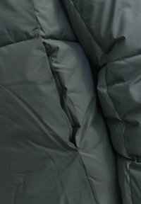 Dunkelgrüner Pufferjackenstoff mit sichtbaren Nähten, plüschiger Textur und gestepptem Muster. Verfügt über eine Seitentasche und eine glatte Oberfläche.